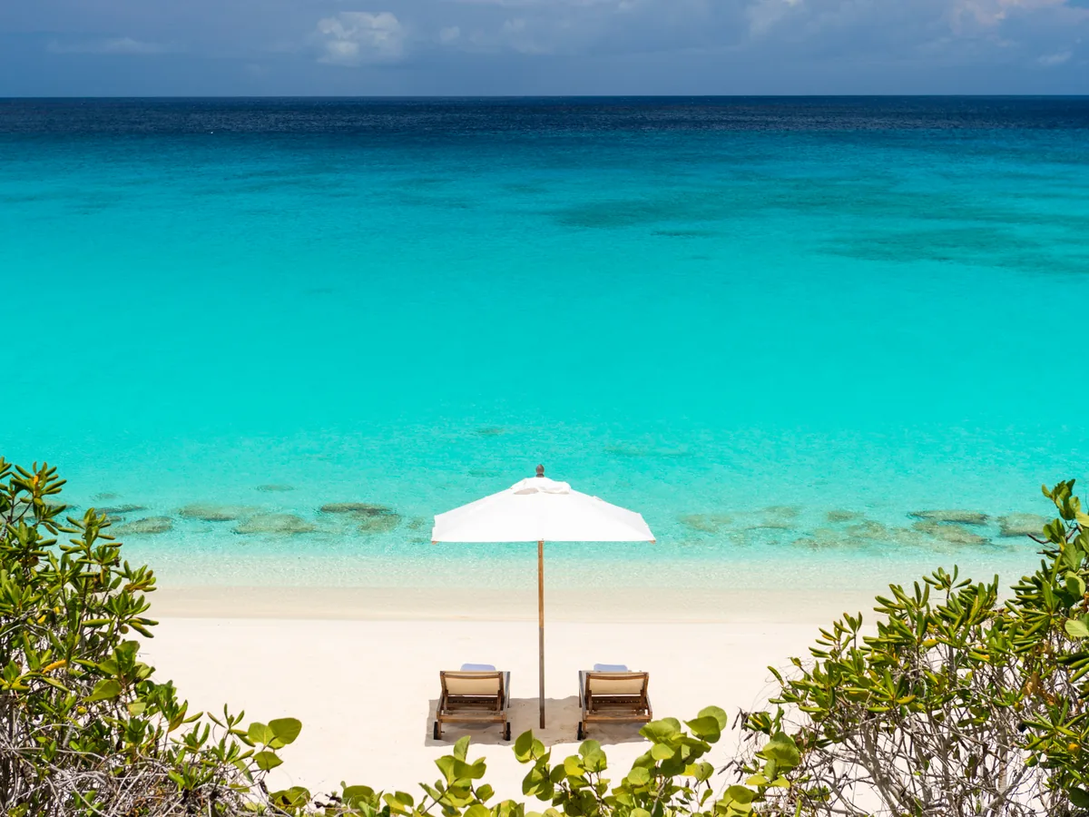 A single cream umbrella and two loungers on white sand above deep turquoise water