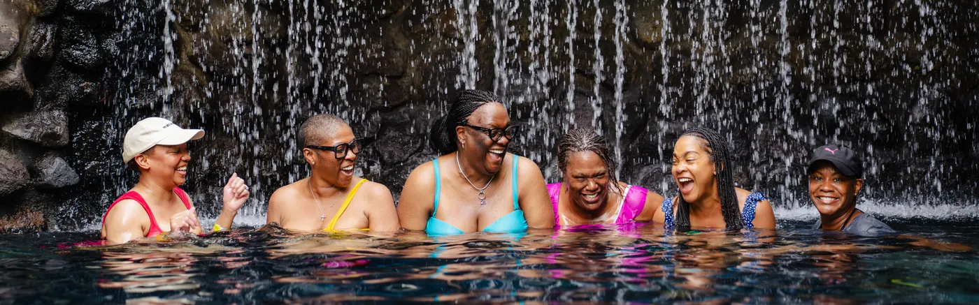 Six women laughing together in a dark pool beneath a stone waterfall