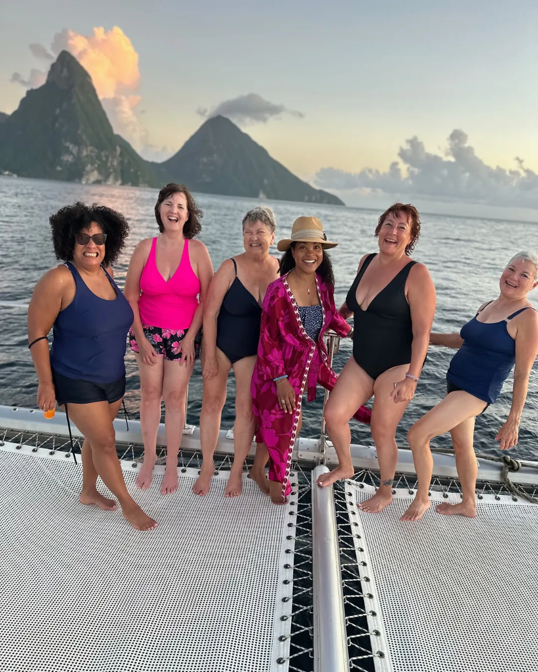 Six women on the bow of a catamaran at golden hour, the Pitons of Saint Lucia rising behind them